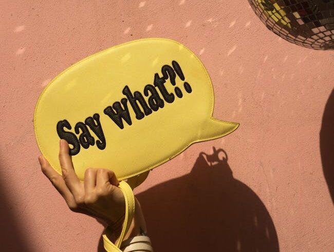 Uma mão segurando um balão de diálogo amarelo com o texto “O quê?!” contra uma parede rosa.